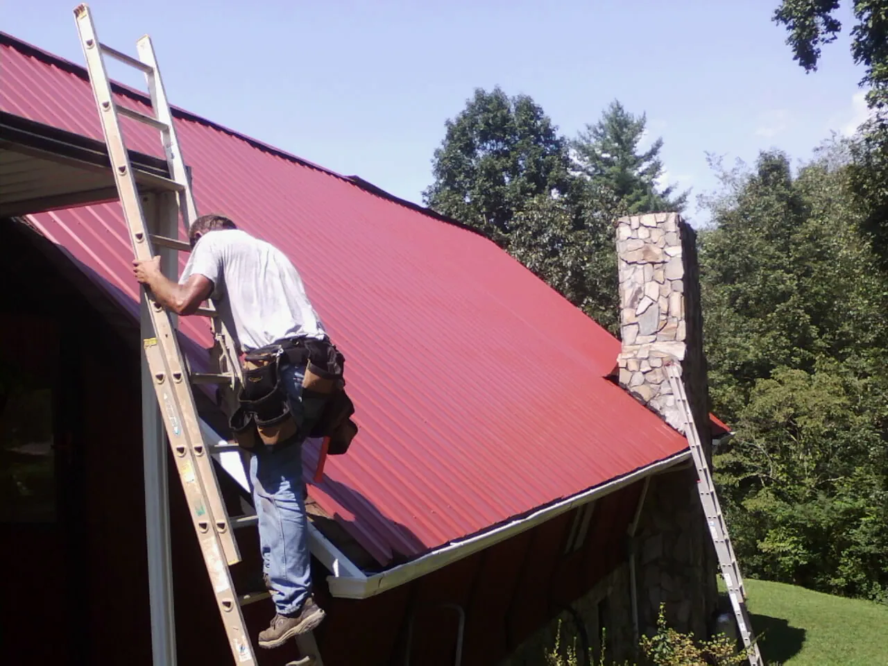 Roofer on a ladder beside a red metal roof and stone chimney — Soffit & Fascia Repair by Roofing Brothers in Tidioute